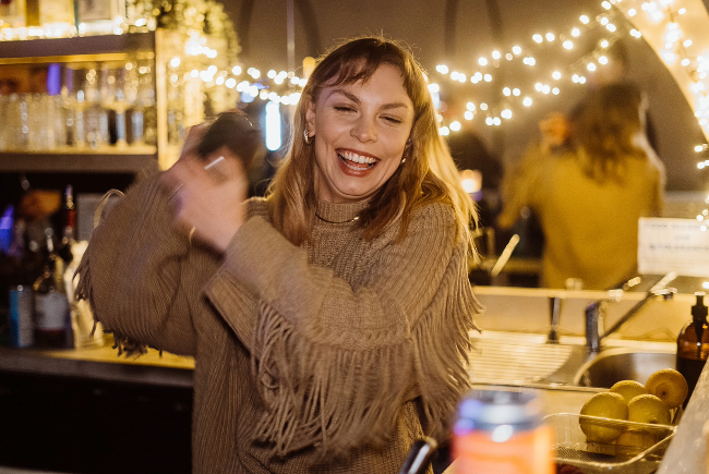 Smiling barwoman shaking a cocktail in a shaker over her shoulder.